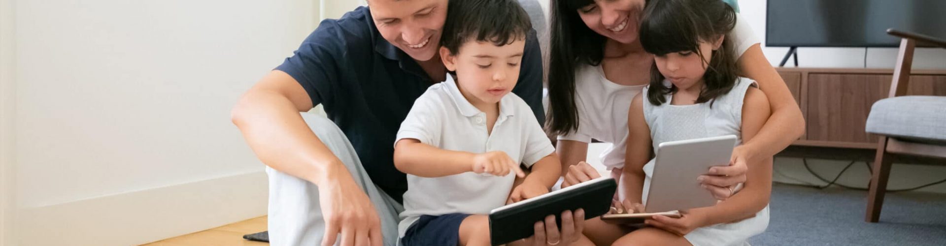 Happy parents and cute kids using mobile devices on floor in living room. Pretty mother and father hugging children with tablets and smartphone and smiling. Family and digital technology concept